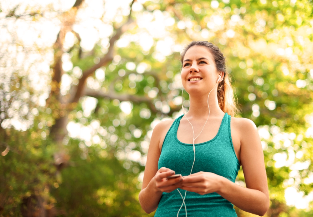 Lächelnde Frau mit Kopfhörern im Grünen – Qnect Walks Audio-Wanderkarten für Ruhe, Verbindung und Klarheit.